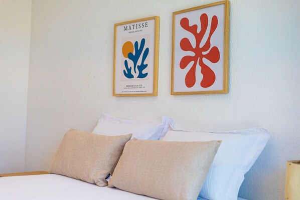A neatly made bed with beige and white pillows, two framed artworks on the wall above it, and a plain white wall.