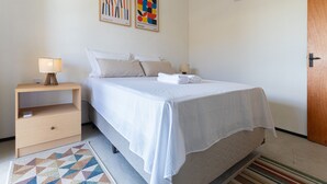 A neatly made bed with a white bedspread and pillows, a wooden nightstand with a lamp, a geometric-patterned rug, and two abstract paintings on the wall. - Beachfront Duplex #a10 in Barro Preto by Carpediem (Aquiraz)