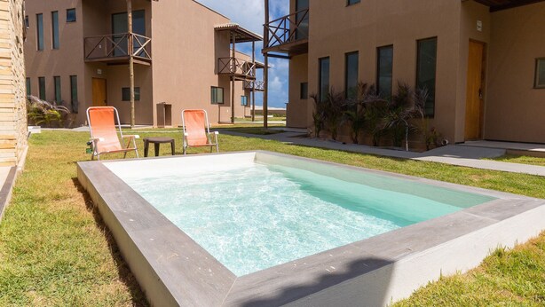 A swimming pool surrounded by a grassy area, with a building featuring balconies and a door. - Beachfront Duplex #a10 in Barro Preto by Carpediem (Aquiraz)