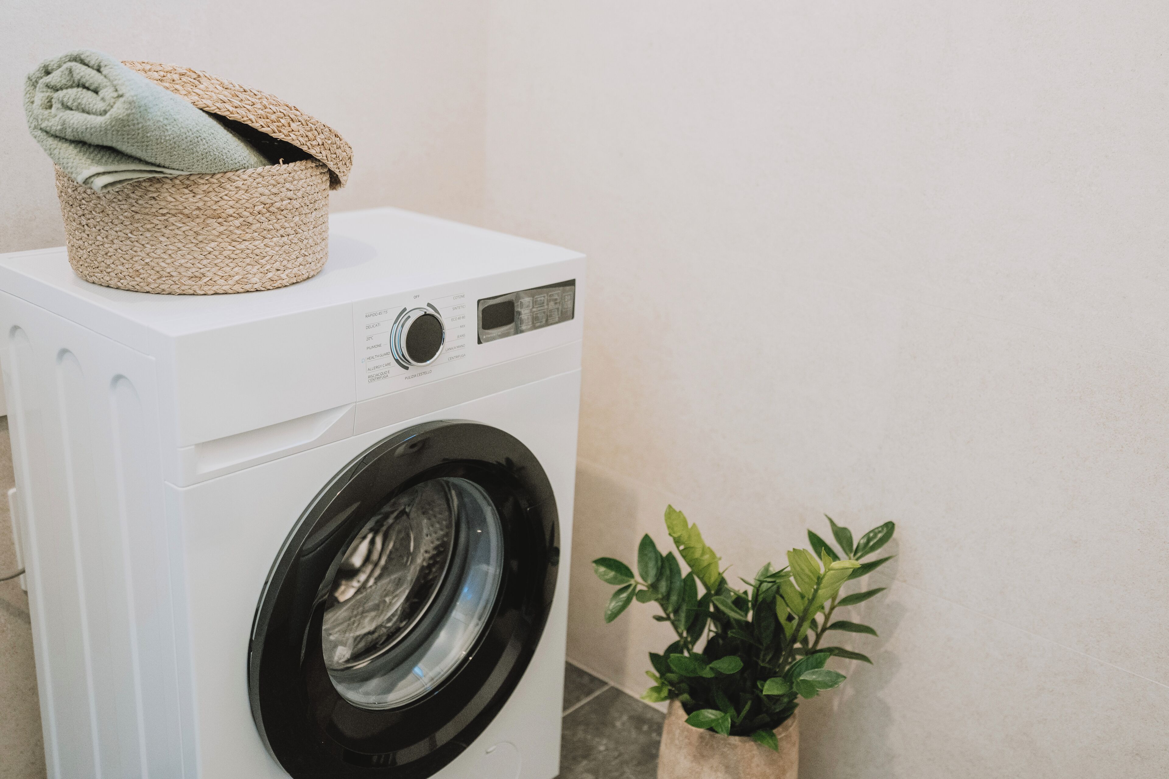 Een wasmachine met een voorlaadklep, met een mandje erbovenop, een bloempot ernaast en een effen muur op de achtergrond.