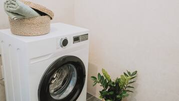 Een wasmachine met een voorlaadklep, met een mandje erbovenop, een bloempot ernaast en een effen muur op de achtergrond.