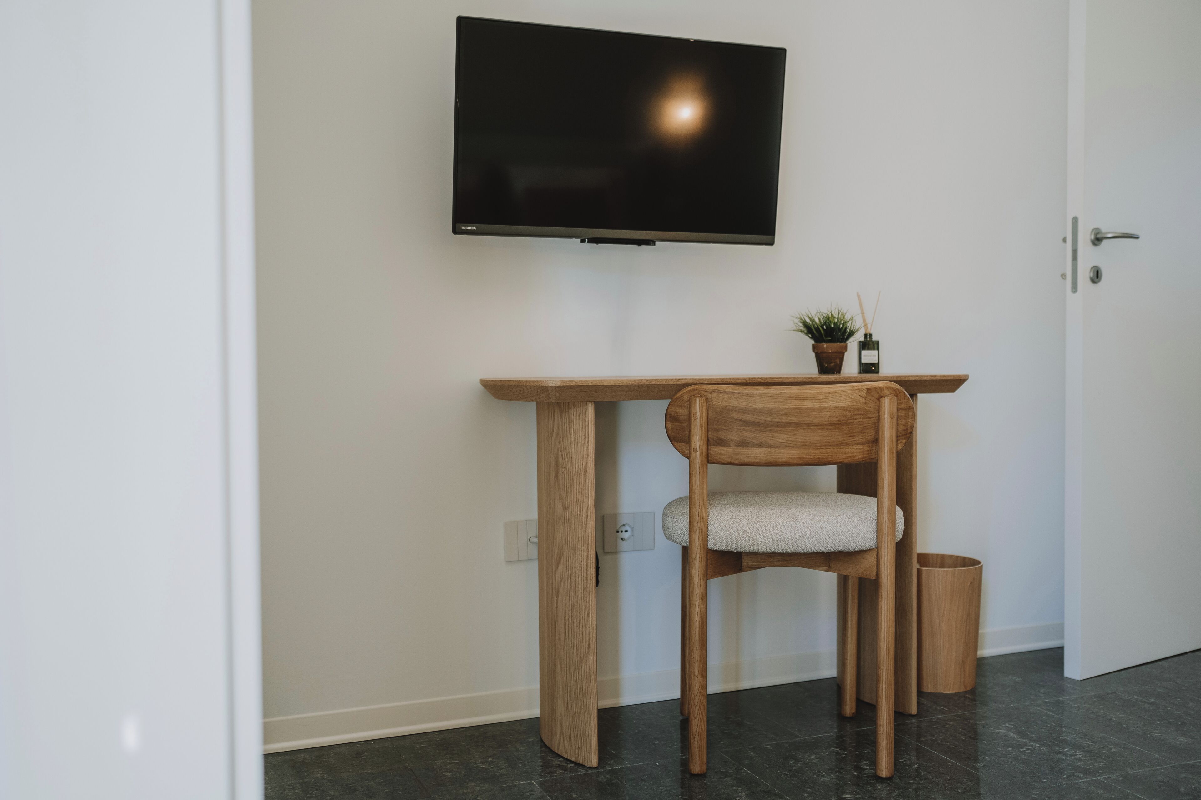 A room with a TV mounted on the wall, a wooden desk with a chair, a small potted plant, and a trash bin.