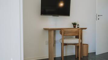 A room with a TV mounted on the wall, a wooden desk with a chair, a small potted plant, and a trash bin.