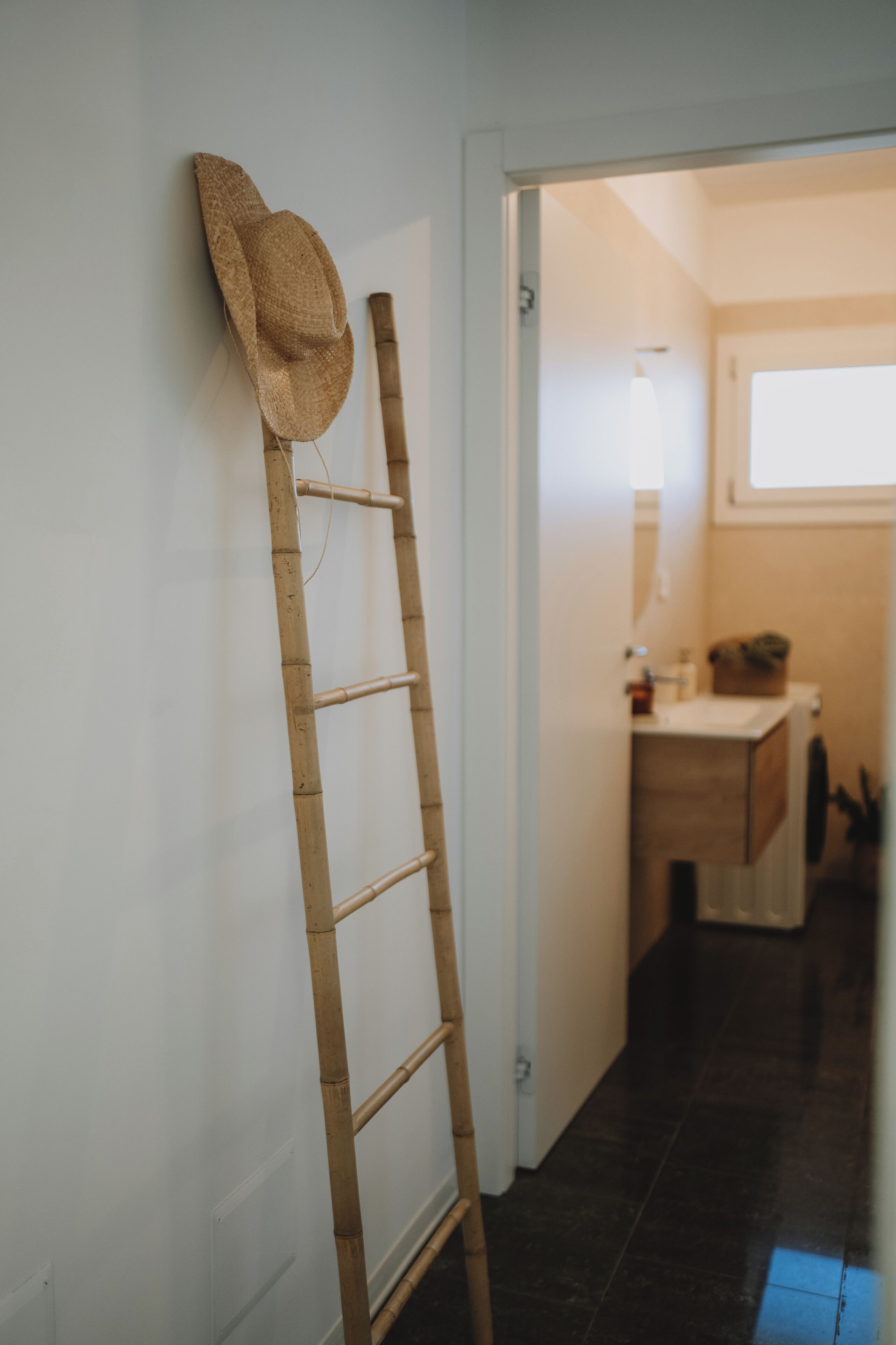 Een houten ladder die tegen een muur staat, met een strooien hoed bovenop, leidend naar een kamer met een raam en een wastafel.