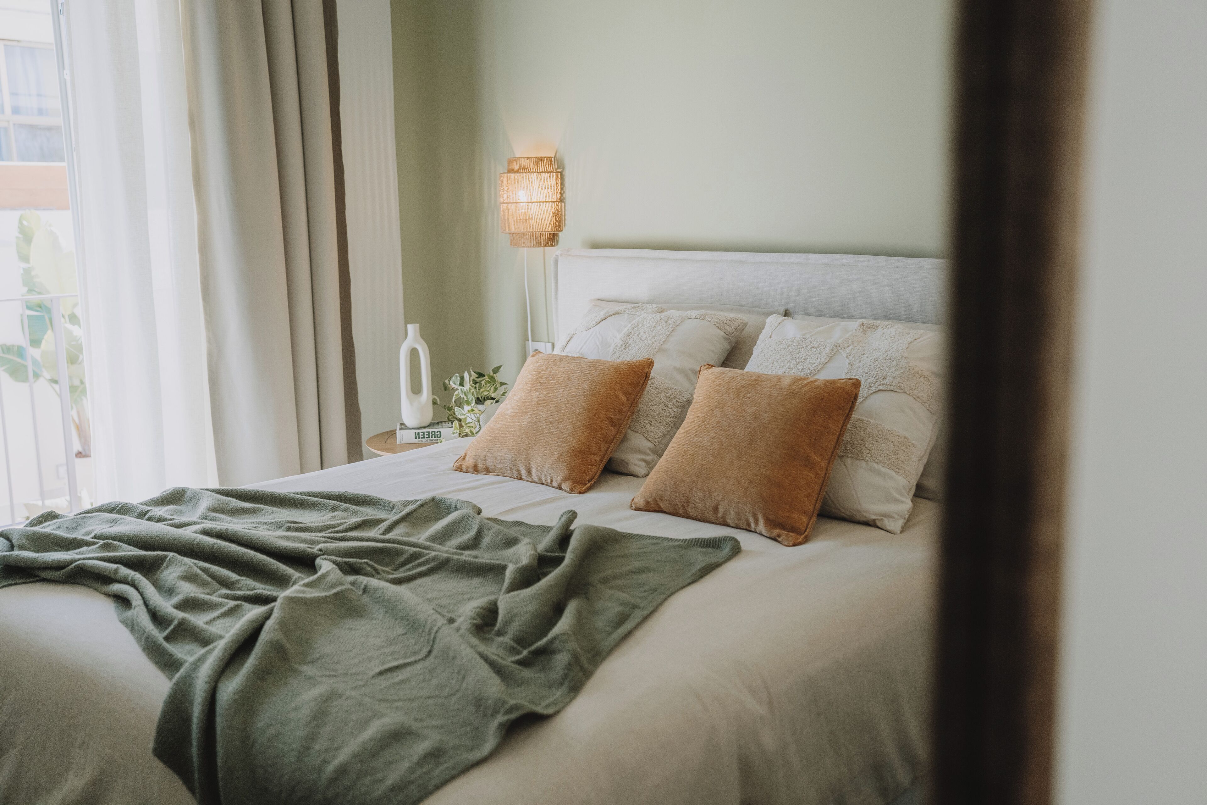 Een netjes opgemaakt bed met een groene deken, een bedlampje en een nachtkastje met een witte vaas en een plant.