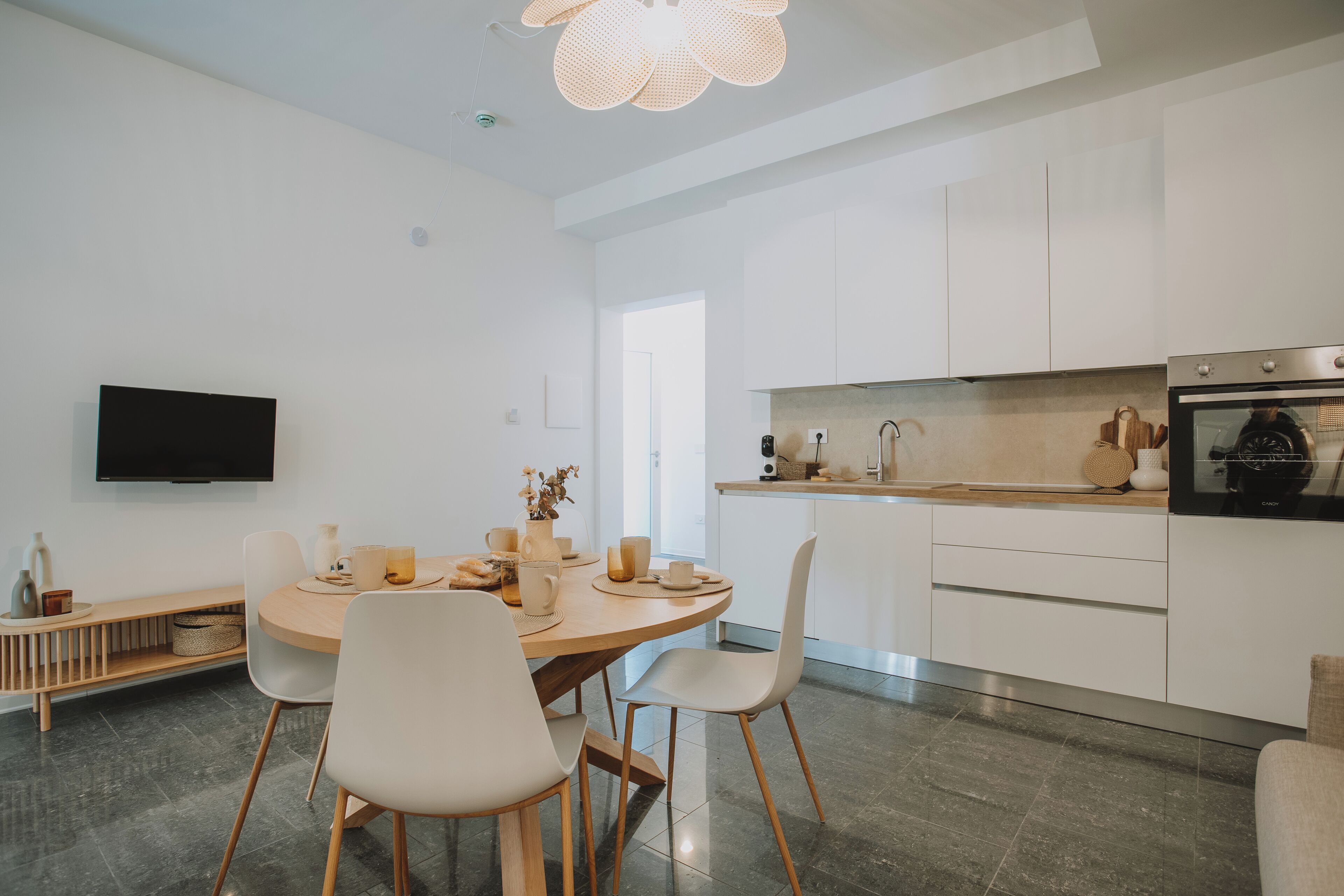 Una cocina moderna con armarios blancos, un comedor con una mesa redonda y sillas, un televisor de pantalla plana en la pared y una lámpara en el techo.