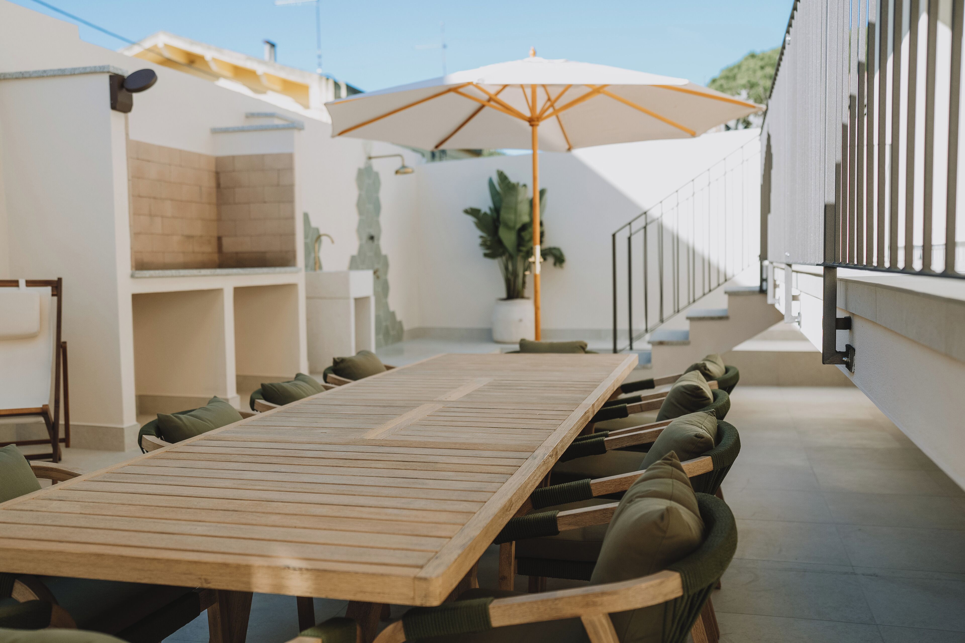Een lange houten tafel met groene stoelen met kussens, een witte parasol en een trap naar een hoger gelegen niveau.