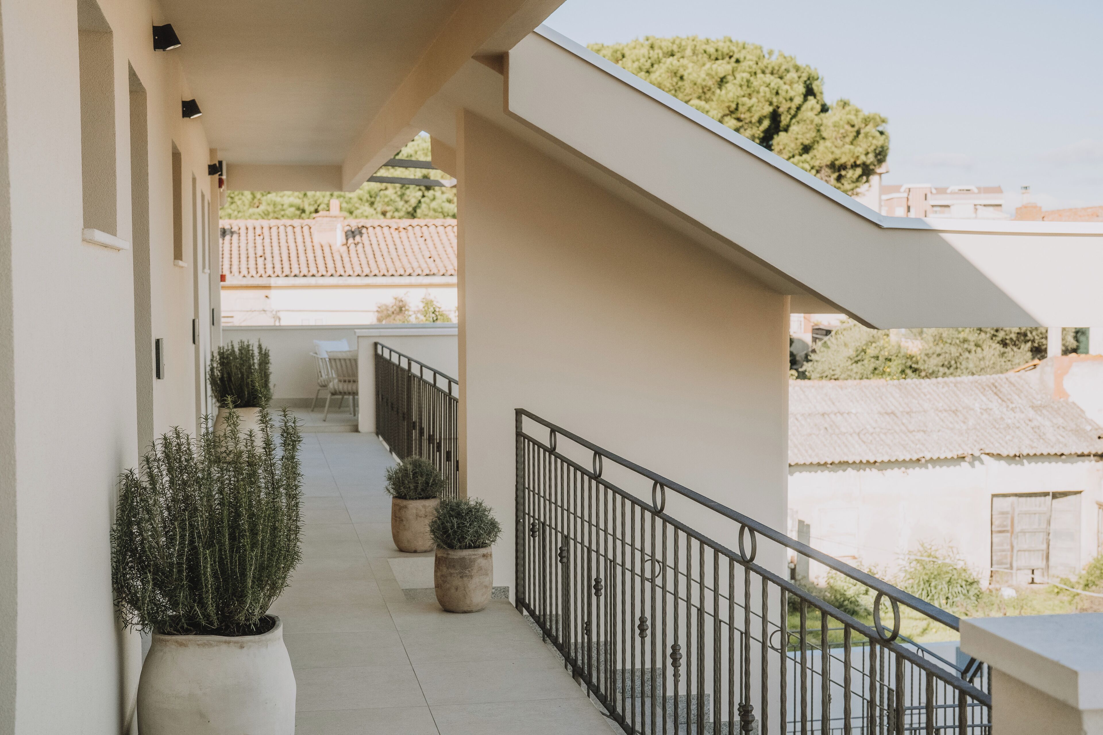 A covered balcony with potted plants, a railing, and a view of buildings and trees.