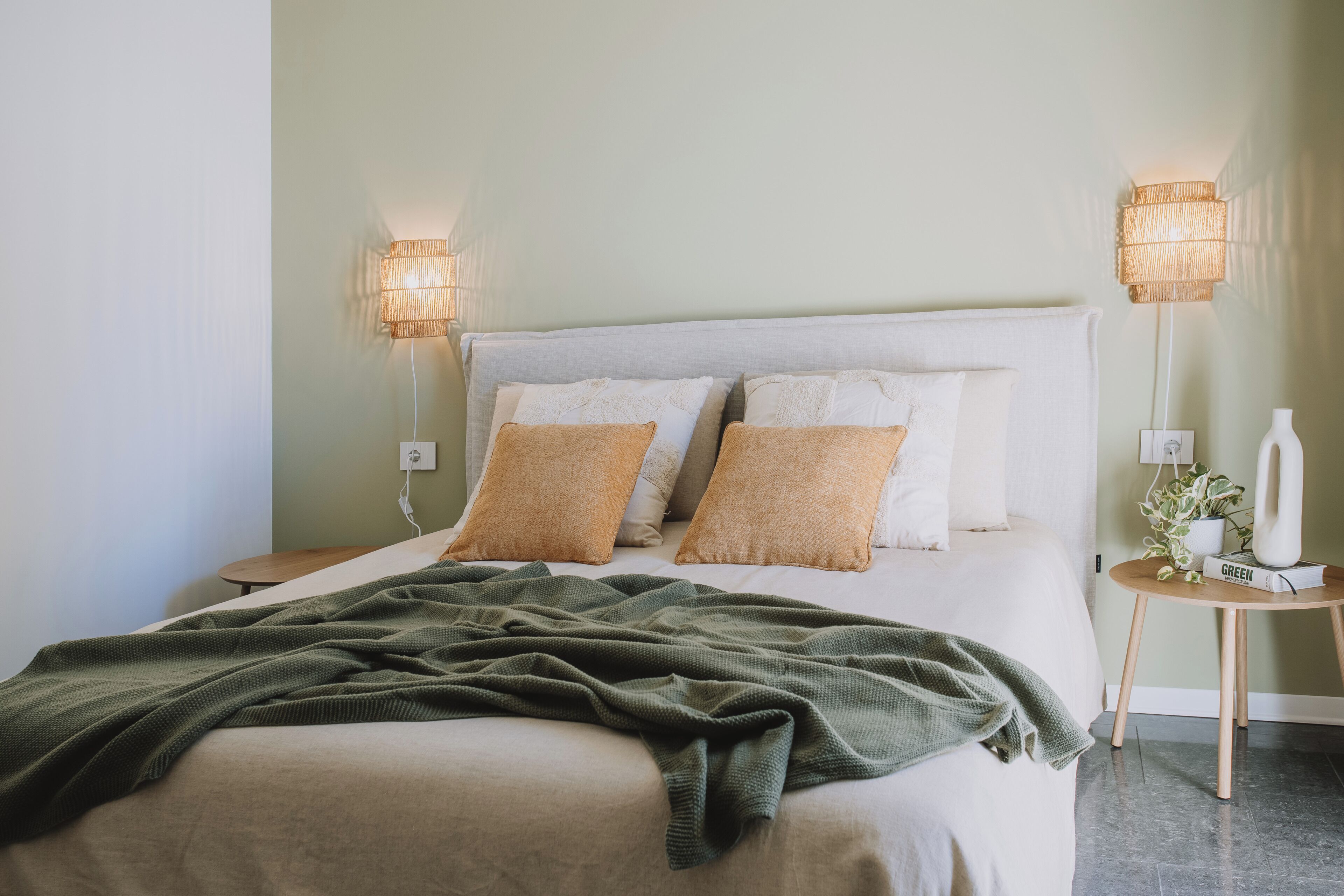 A neatly made bed with a green blanket, two bedside lamps, and a small bedside table with a plant and a book.