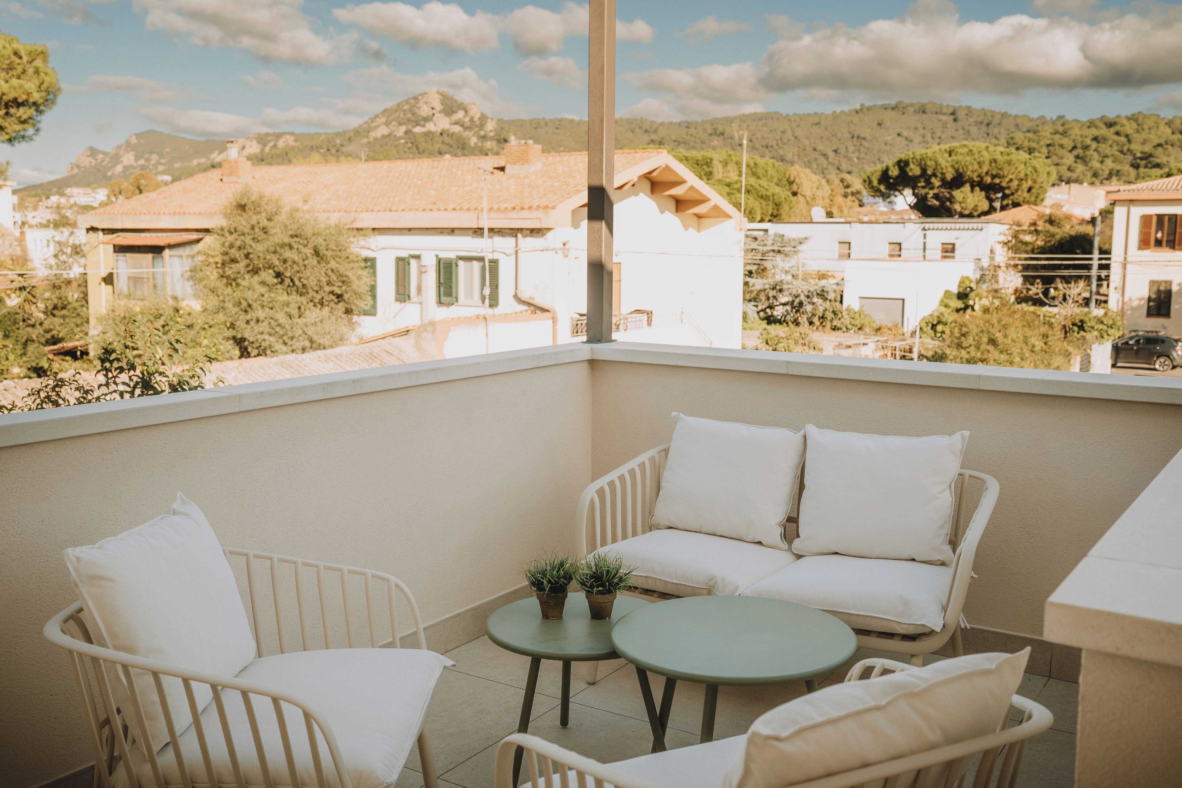A balcony with white furniture, two potted plants, and a view of buildings and hills.
