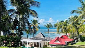Un resort frente al mar con una zona con carpas blancas y un edificio de techo rojo, rodeado de palmeras y el océano.