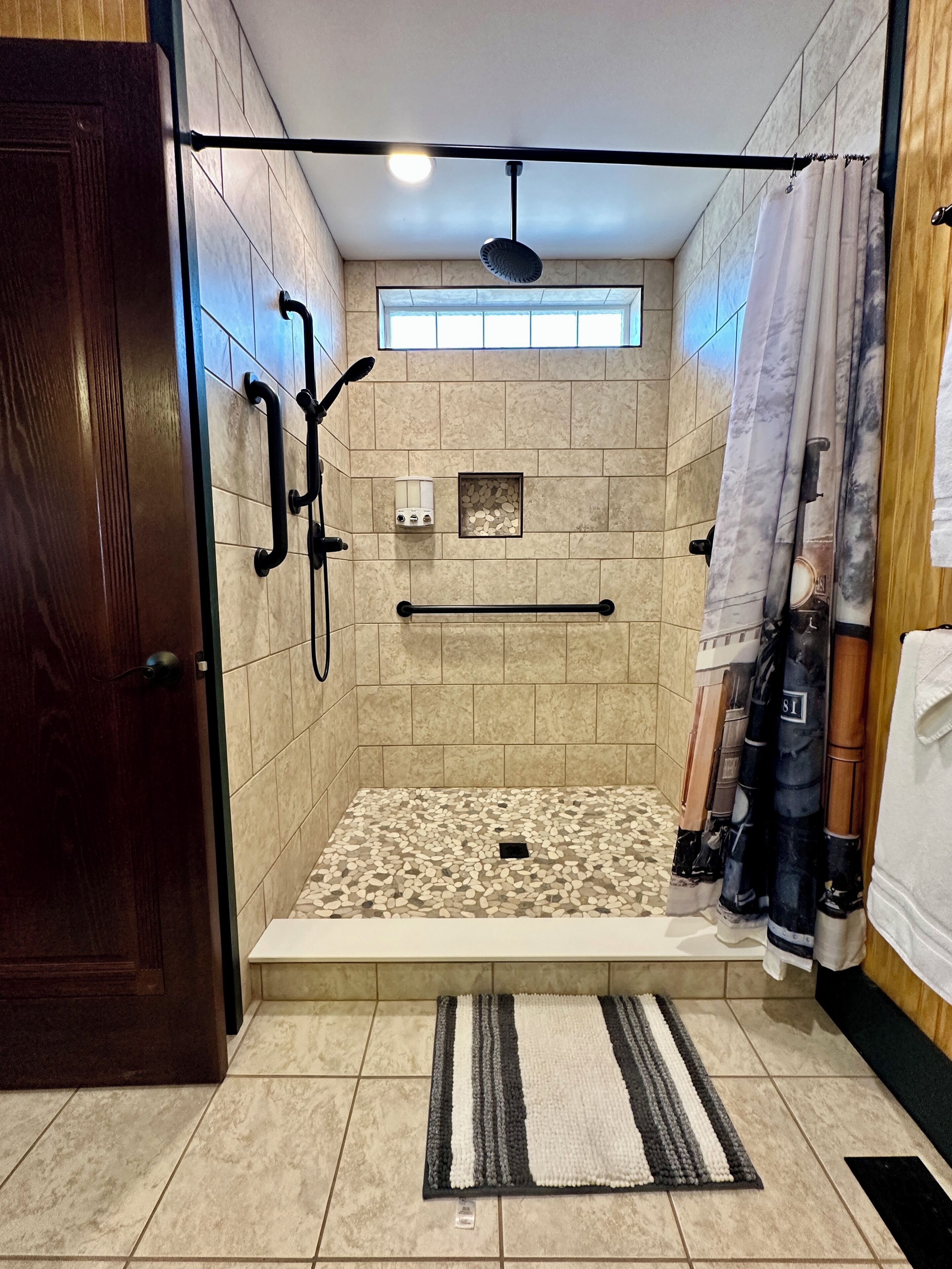 A walk-in shower with a tiled floor, a handrail, a ceiling fan, and a window.