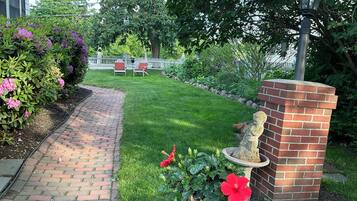 A brick pathway leading to a white picket fence, a statue, and a garden with various plants.