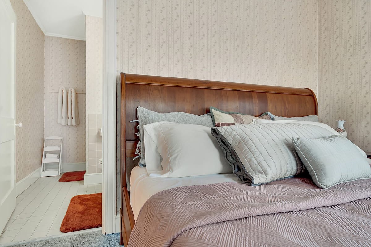 A bedroom with a wooden headboard, a bed with multiple pillows, a white bedside table, and a red rug.