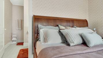 Un dormitorio con cabecera de madera, cama con múltiples almohadas, mesita de noche blanca y alfombra roja.