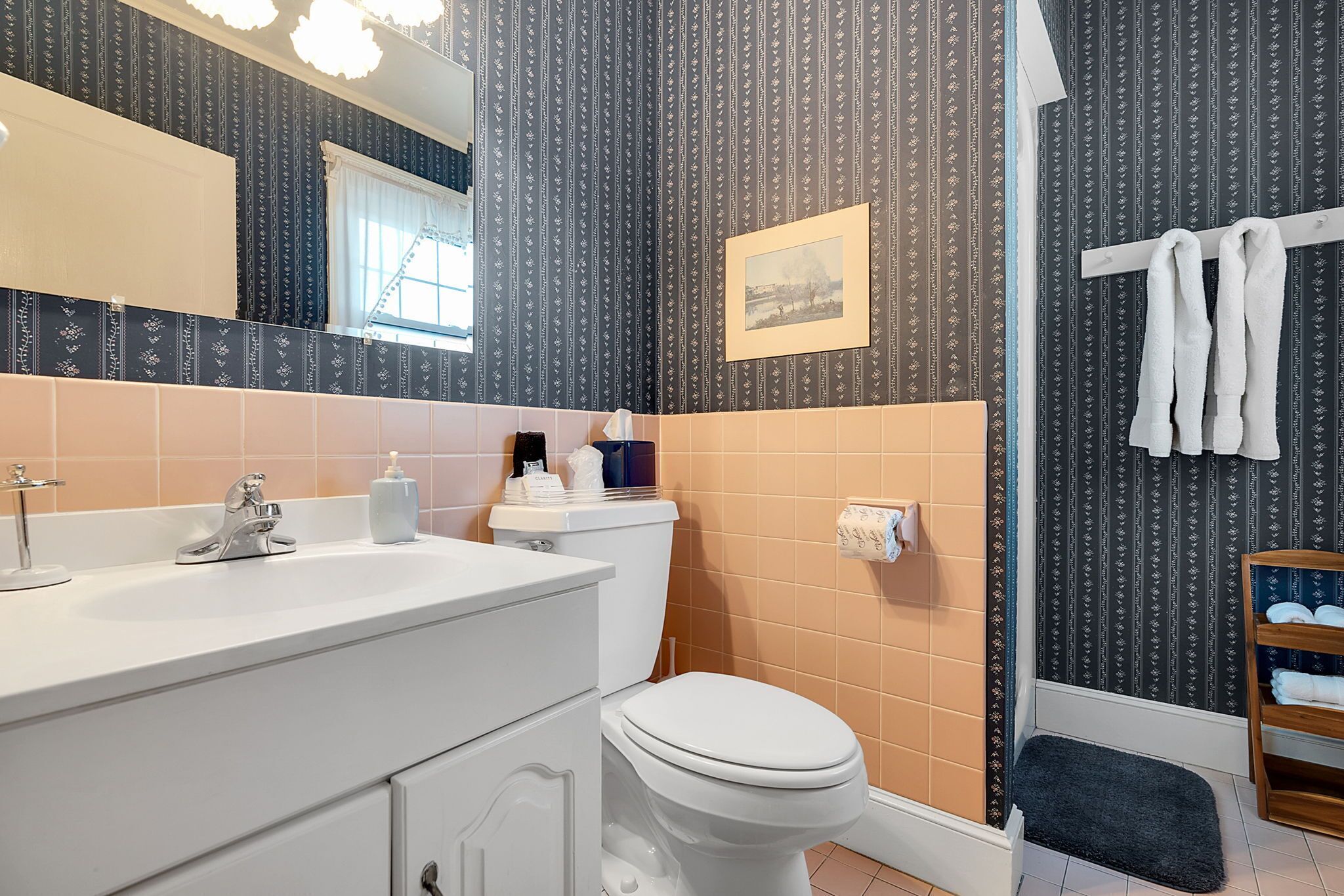 Een badkamer met een wit wastafelmeubel, toilet en douchegedeelte. Handdoekrek met handdoeken, een foto aan de muur en een klein plankje met toiletartikelen.