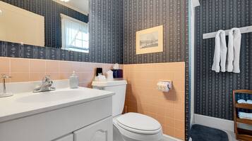 Een badkamer met een wit wastafelmeubel, toilet en douchegedeelte. Handdoekrek met handdoeken, een foto aan de muur en een klein plankje met toiletartikelen.