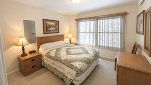 Quarto com cama, duas mesas de cabeceira com luminárias, cabeceira de madeira, janela com persianas e uma escrivaninha com cadeira.