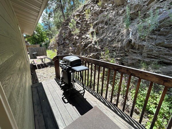 A wooden deck with a grill and a table, surrounded by a rocky cliff and greenery.
