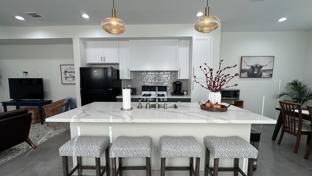 A modern kitchen with a central island, bar stools, a flat-screen TV, and a dining area with chairs and a table.