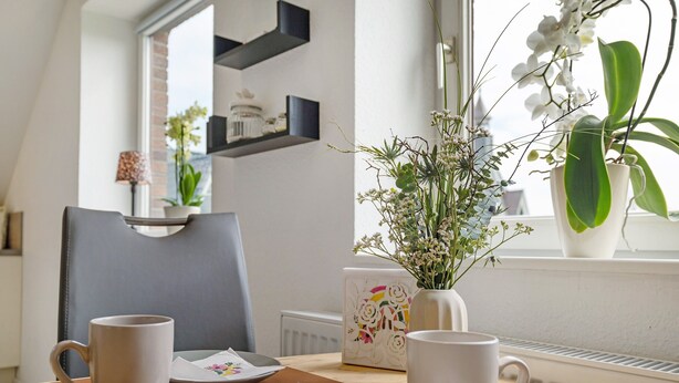 Uma área de refeições com uma mesa posta para dois, um vaso com flores e uma janela com plantas.