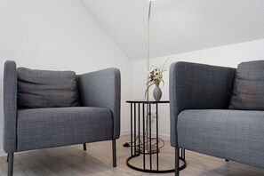A room with two grey upholstered chairs, a small round table with a vase of flowers, and a tall floor lamp.