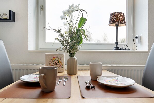 A dining table with two plates, cutlery, and two mugs. There is a vase with flowers and a lamp on the table.
