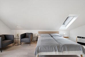 A modern bedroom with a skylight, a bed with a grey bedspread, two grey sofas, a small round table with a lamp, and a bedside table with a lamp.