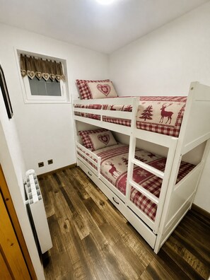 Una cama litera con colcha a cuadros y almohadas con forma de corazón, una ventana con cortina y suelo de madera.