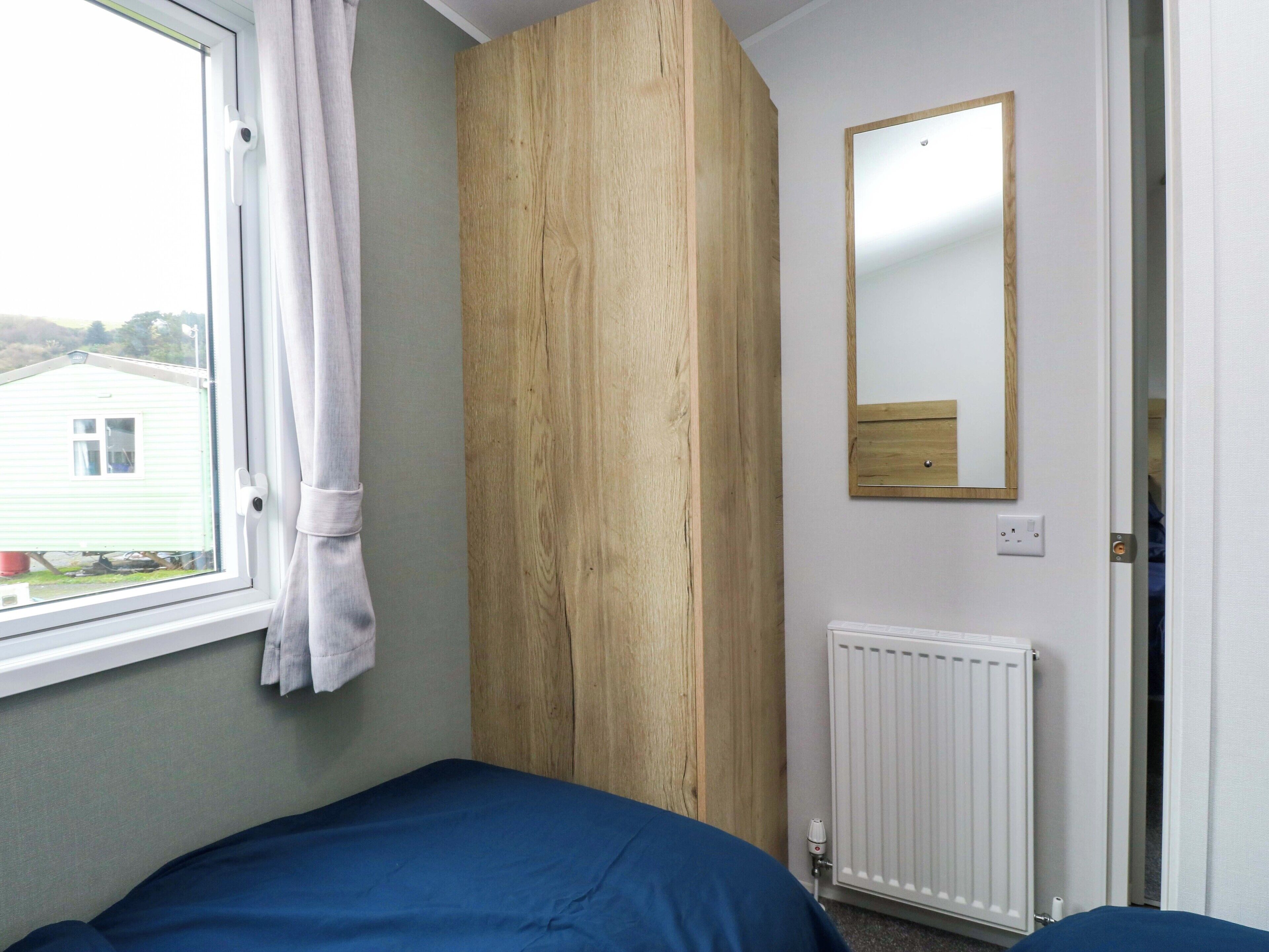 A bedroom with a single bed, a window with curtains, a wooden panel wall, a mirror, and a radiator.