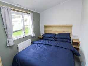 A bedroom with a wooden headboard, a bed with blue bedding, a window with curtains, and a radiator. - The Foxy Cobbler (Kingsbridge)