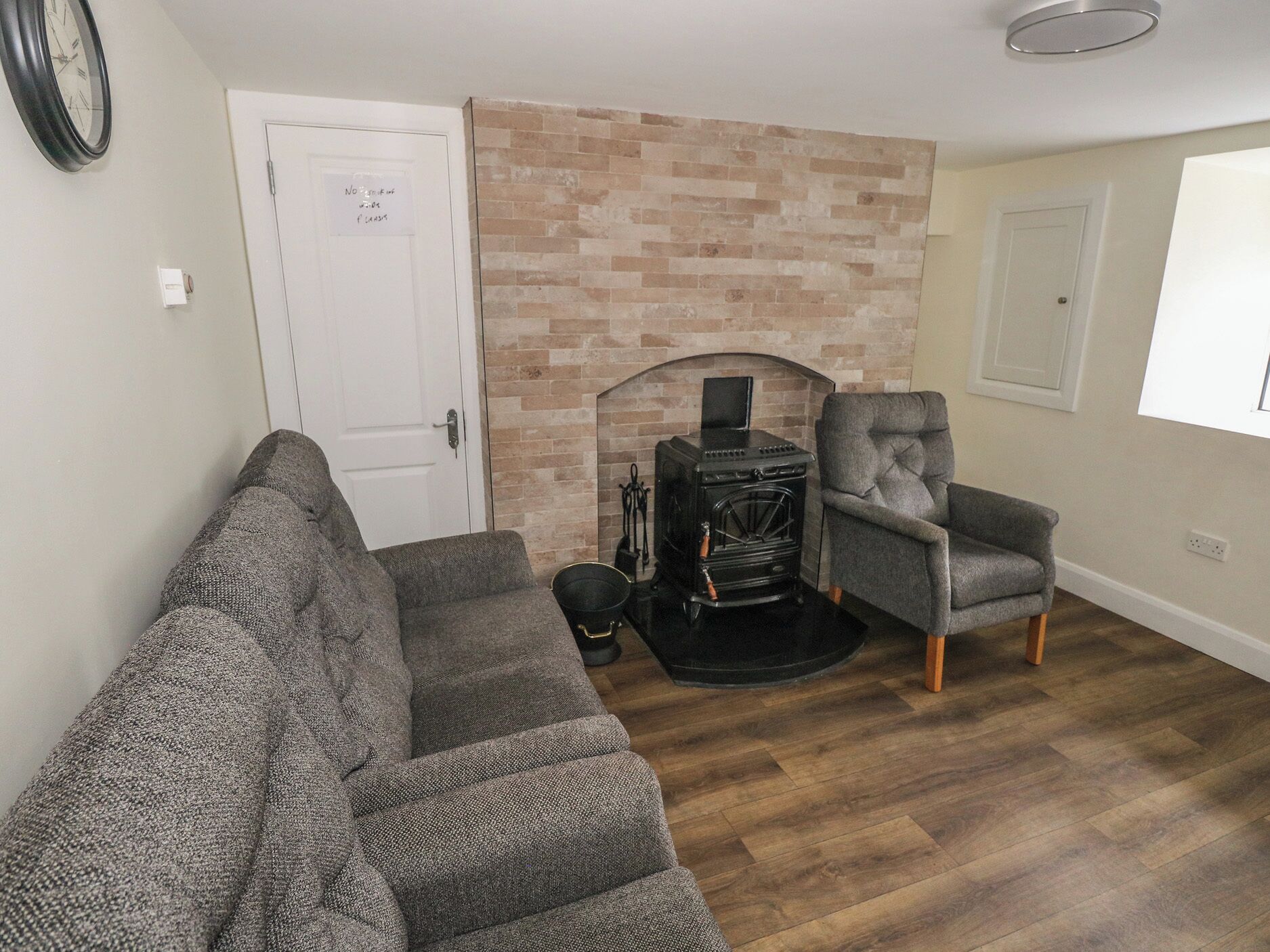 A living room with a brick fireplace, a sofa, a chair, and a clock on the wall.