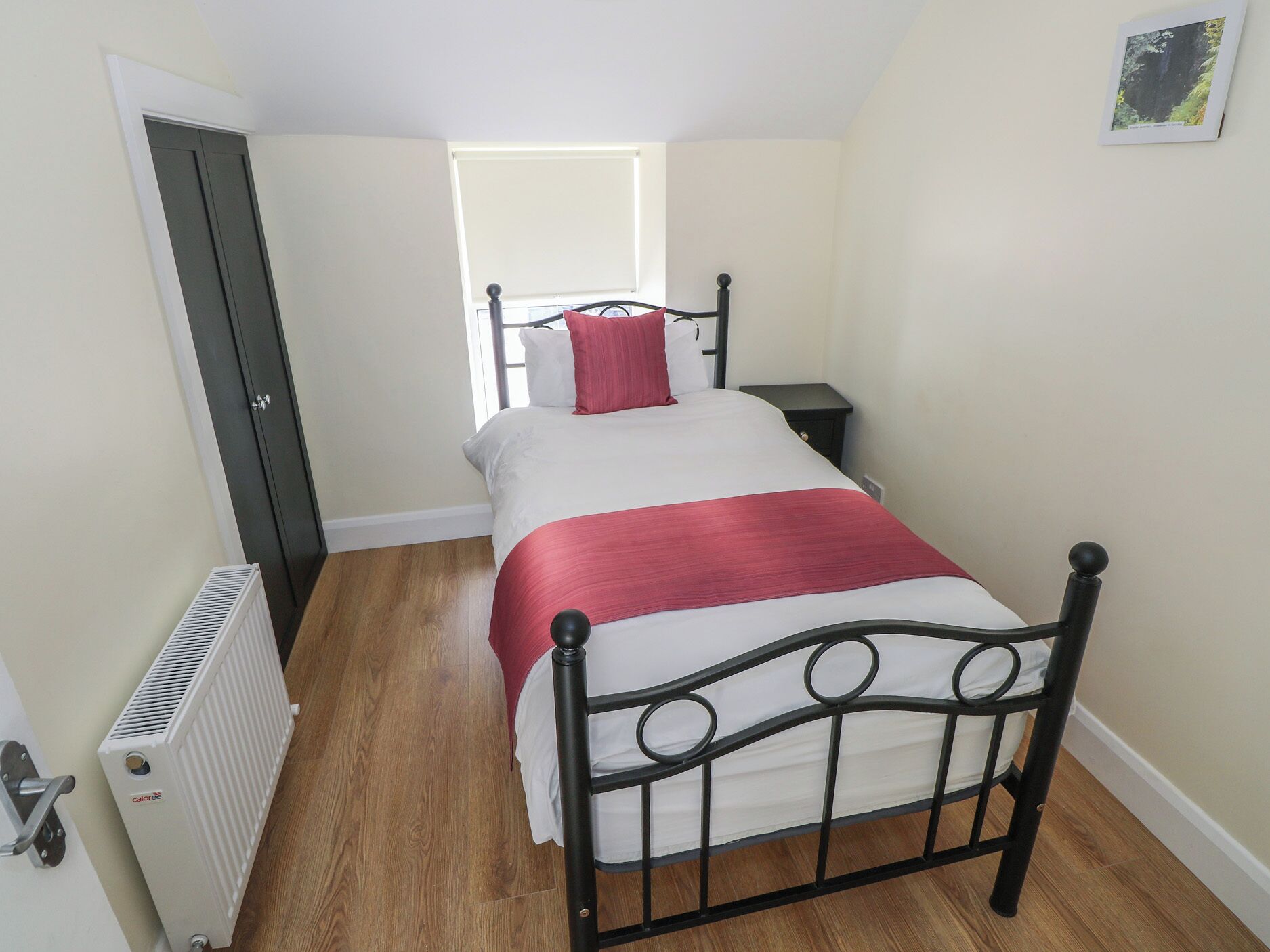 A small, narrow room with a single bed, a bedside table, and a radiator.