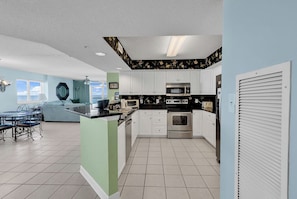 A modern kitchen with white cabinets, stainless steel appliances, and a dining area with a blue couch and table. - Oceanfront 4BR/3BA Luxury Condo in the Laguna Keys in Cherry Grove (North Myrtle Beach)