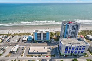 Eine Küstenstadtansicht mit Strand, Parkplatz und mehreren Gebäuden, darunter ein Hotel mit blau-weißer Fassade.
