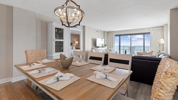 A dining area with a wooden table set for four, a chandelier, and a view of the balcony. - Oceanfront Luxury 3BR/2BA  End Unit with Pool, Fitness Room and Hot Tubs. (North Myrtle Beach)