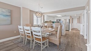 A dining area with a table set for four, white chairs, a chandelier, and a view of the kitchen in the background. - Oceanfront Luxury 3BR/2BA  End Unit with Pool, Fitness Room and Hot Tubs. (North Myrtle Beach)
