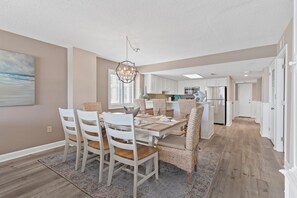 A dining area with a table set for four, white chairs, a chandelier, and a view of the kitchen in the background. - Oceanfront Luxury 3BR/2BA  End Unit with Pool, Fitness Room and Hot Tubs. (North Myrtle Beach)