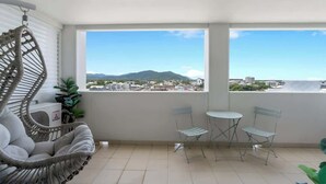 Un balcon avec vue sur la ville et les montagnes, une chaise en osier, deux chaises en métal et une petite table.