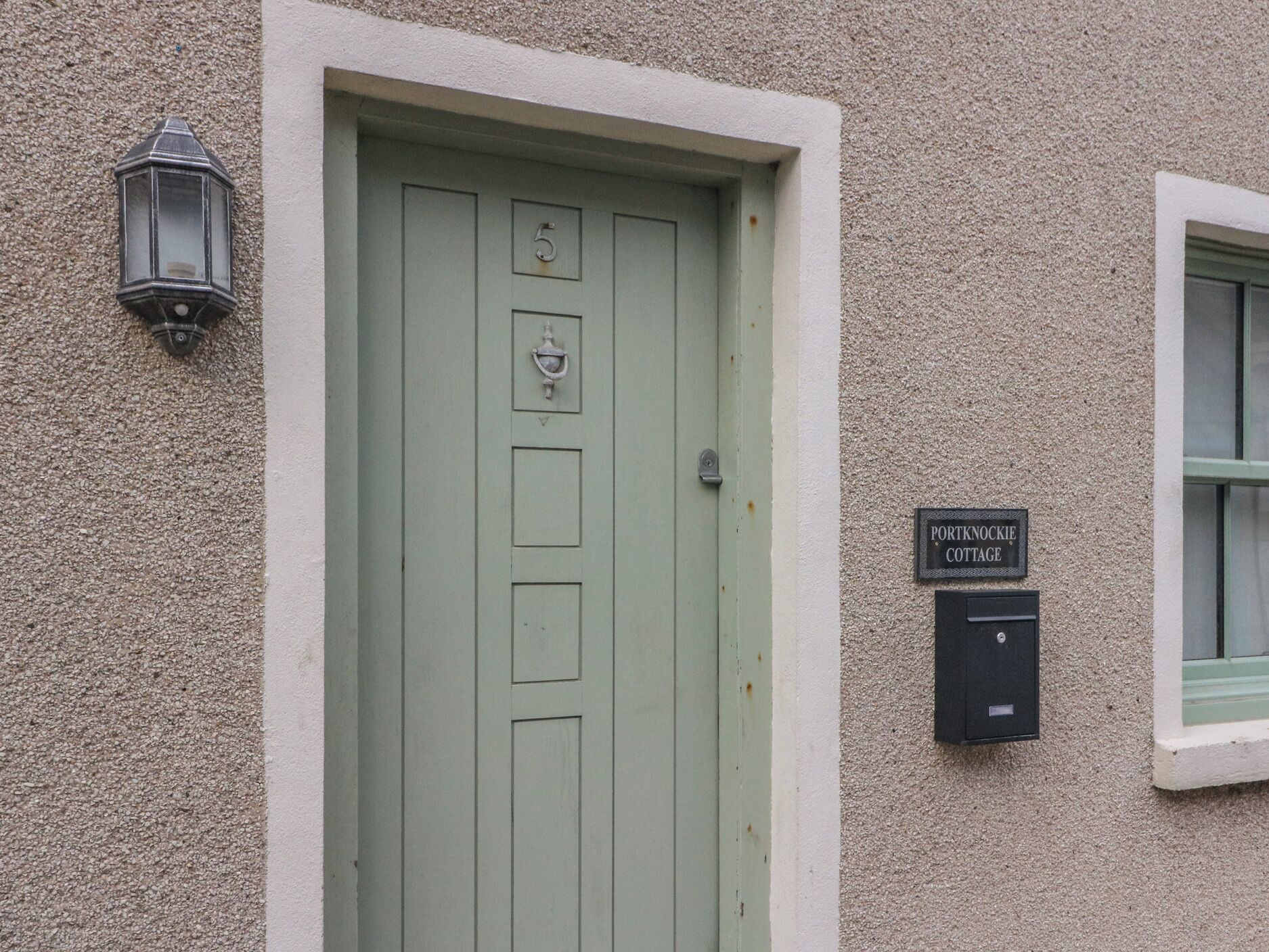 A green door with a number '5' and a doorbell, a mailbox, and a sign reading 'Portknockie Cottage' on a textured wall.