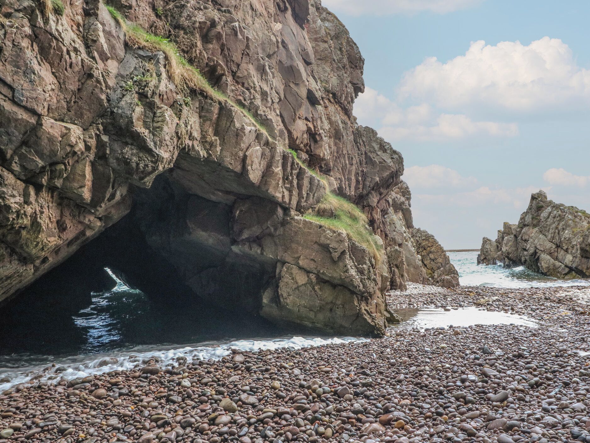 Un paisaje costero rocoso con la entrada de una cueva, una playa de piedras y mar a lo lejos.
