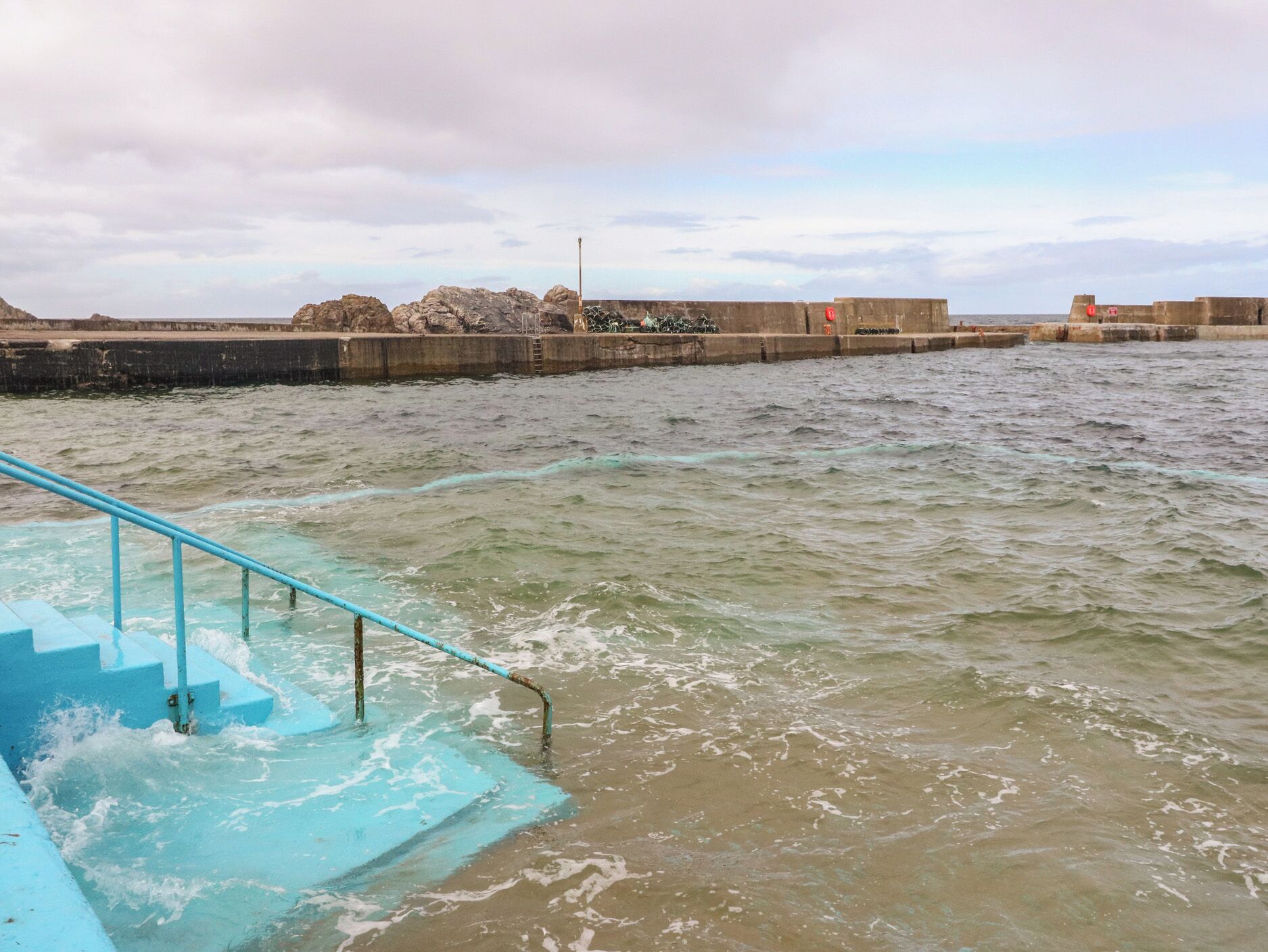 Un paseo marítimo con una escalera azul que desciende al agua, una barrera de hormigón y un cielo nublado.