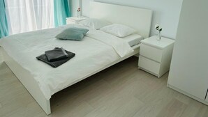 A neatly made bed with white bedding and a folded gray blanket, a white bedside table with a vase of flowers, and a window with sheer white curtains and a teal drape. - Galaxy Residence Villa (Odăile)