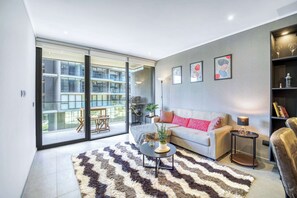 A modern living room with a sofa, a coffee table, and a rug. There is a sliding glass door leading to a balcony with outdoor furniture.