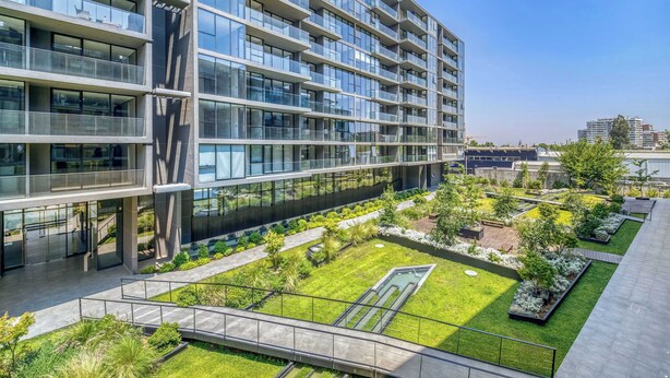 Un edificio moderno de varios pisos con balcones, un jardín en la azotea y un cielo despejado.