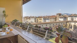Un balcó amb preparació per esmorzar, incloent fruites i suc, amb vista a una ciutat i edificis tradicionals.