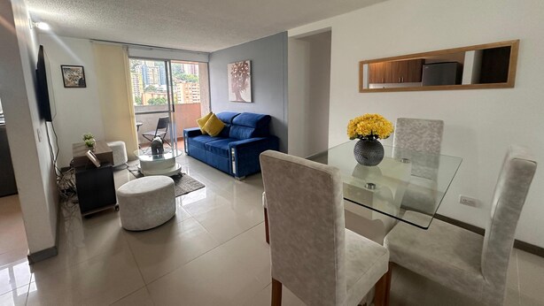 A modern living room with a blue sofa, a glass dining table, and a vase of yellow flowers. - View + Swimming Pool Fast WiFi | 100 Meters subway". (Sabaneta)