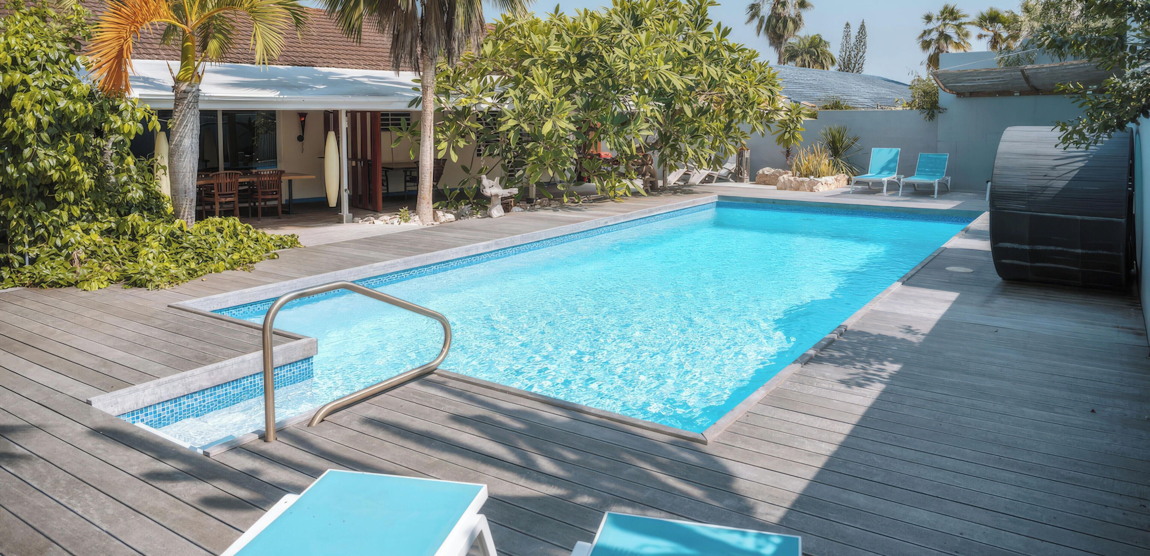 A swimming pool with a wooden deck, lounge chairs, and palm trees.