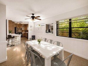 Cozinha moderna com ilha central, sala de jantar com mesa e cadeiras brancas, e uma janela grande com vista para vegetação.