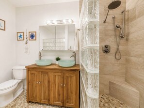Un baño con tocador de madera, dos lavamanos de vidrio, un espejo, ducha con cerramiento de vidrio y un inodoro.